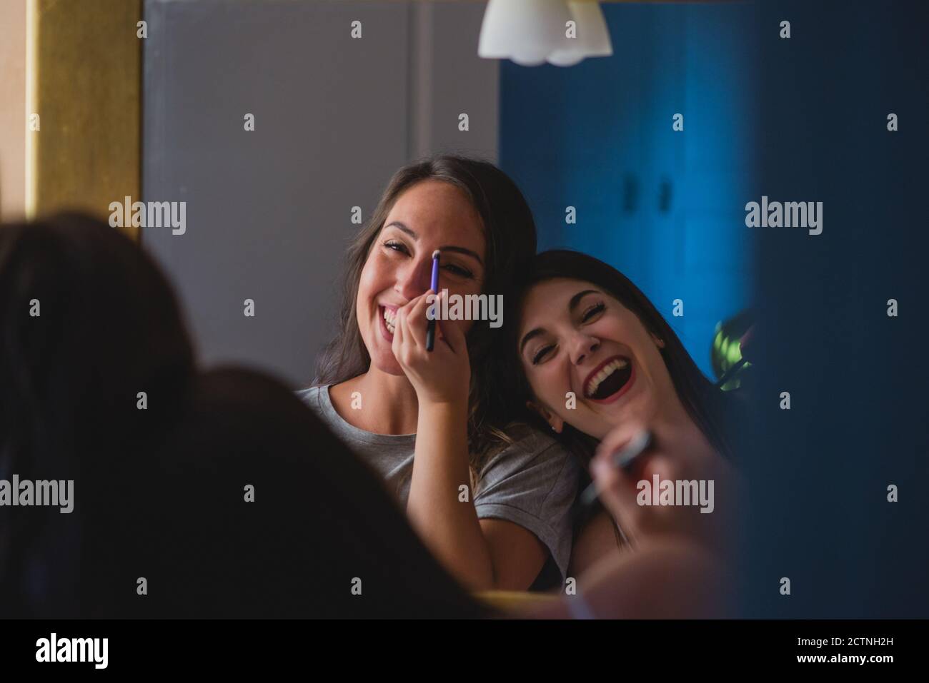 Delighted female friends standing in front of mirror at home and doing makeup together while preparing for party celebration Stock Photo