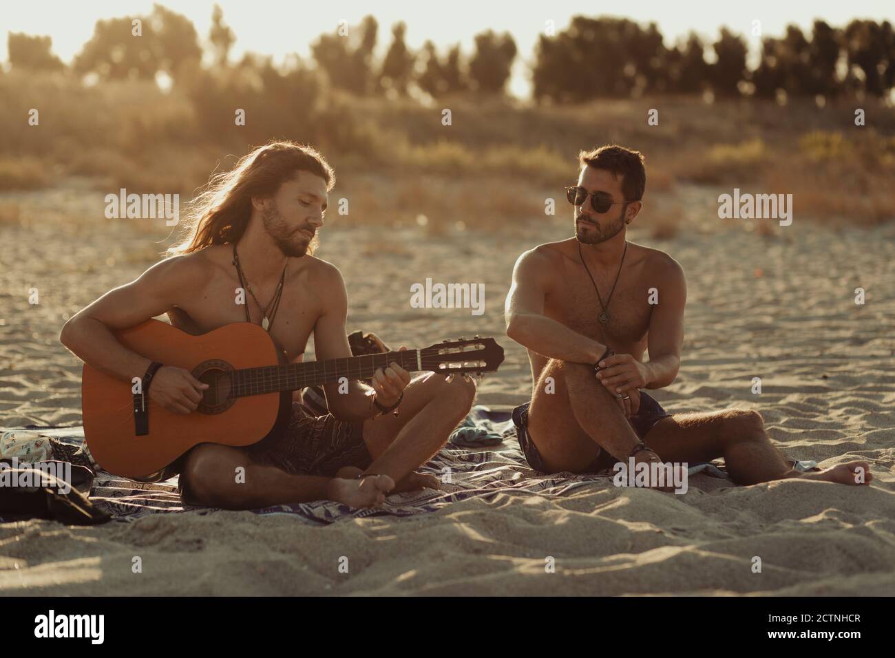 Relaxed talented man playing acoustic guitar for male friend while sitting together on sandy beach at sunset Stock Photo