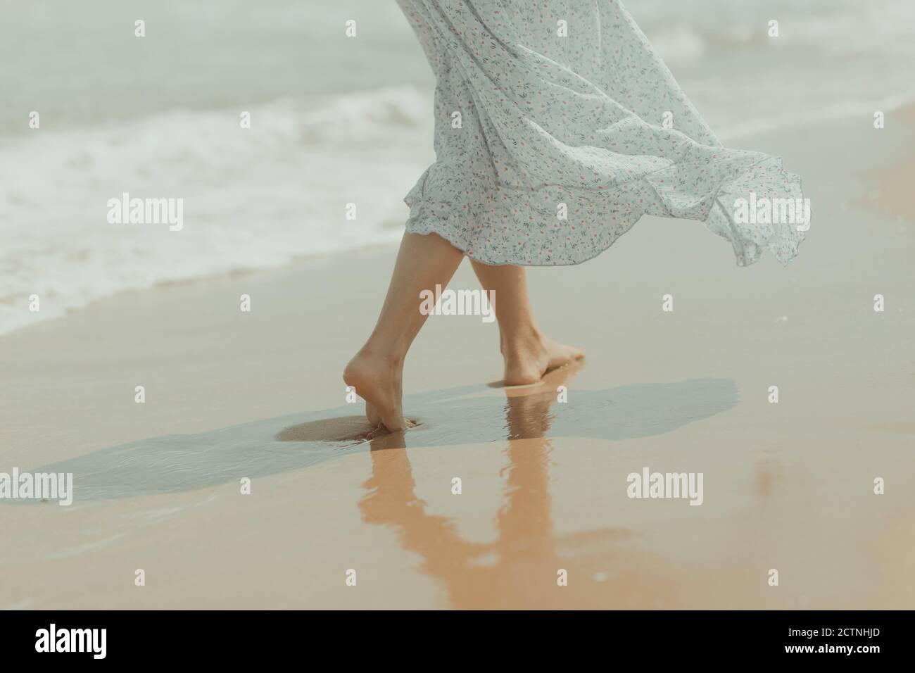 Back view of crop unrecognizable barefoot female in white summer dress walking alone on sandy beach in sunny summer day in Spain Stock Photo