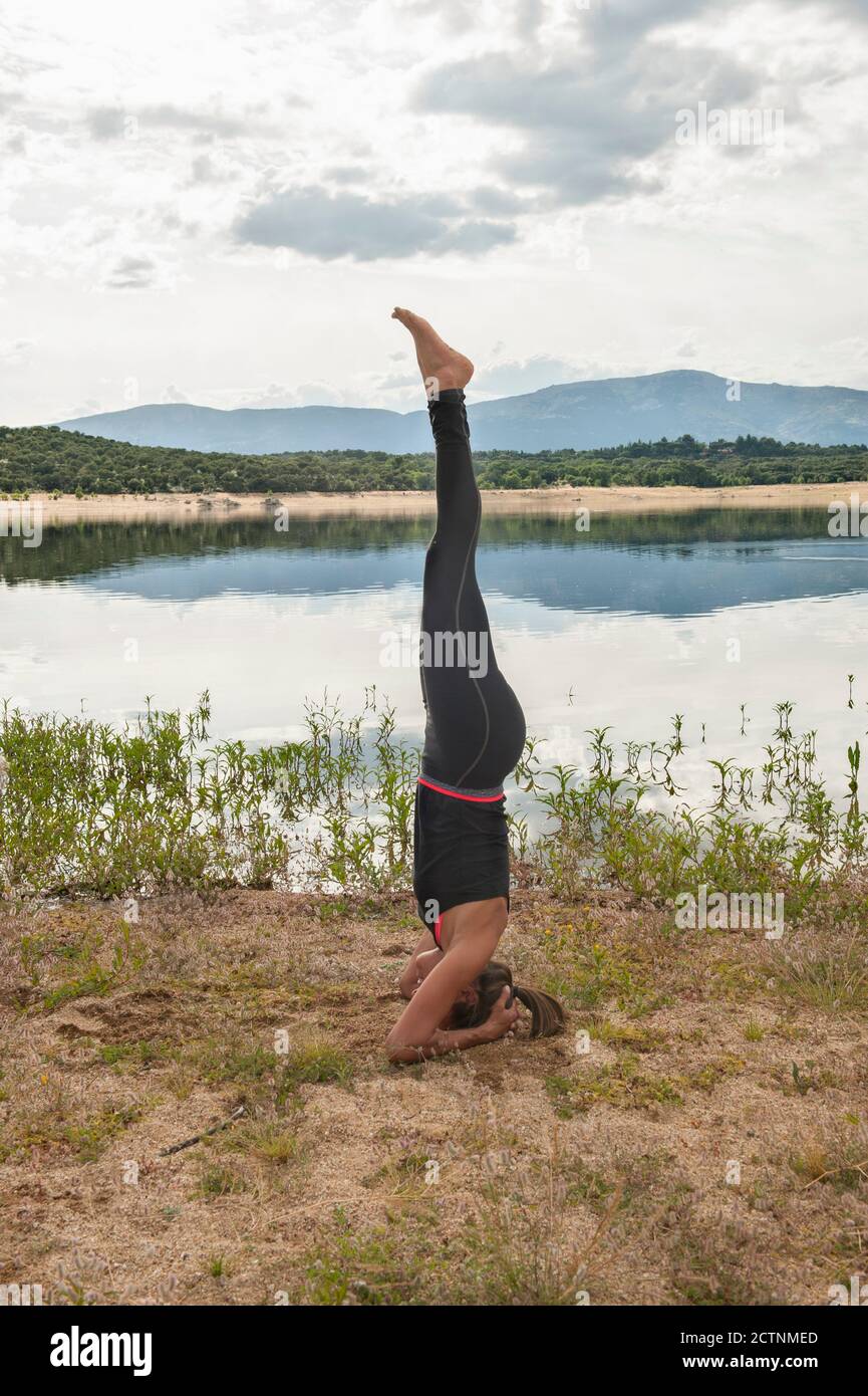 Full body side view of slim female in sportswear doing Salamba Sirsasana yoga pose variation during practice on lake shore Stock Photo