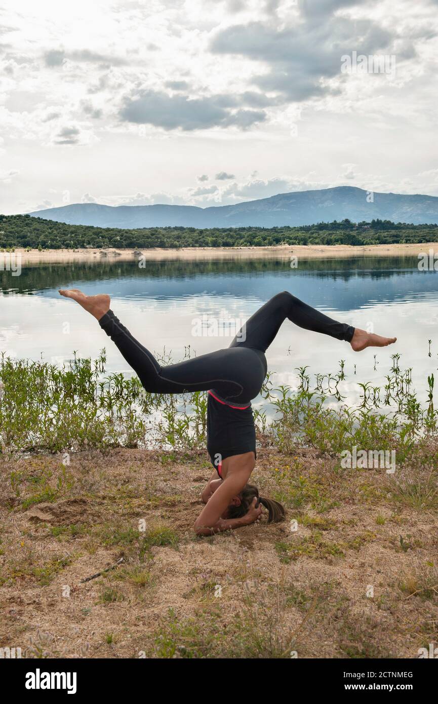 Full body side view of slim female in sportswear doing Salamba Sirsasana yoga pose variation during practice on lake shore Stock Photo