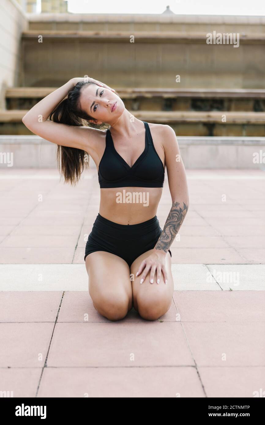 Full body of young fit female in black sports bra and shorts performing  stretching exercise for neck and doing head side bend while working out on  paved square Stock Photo - Alamy