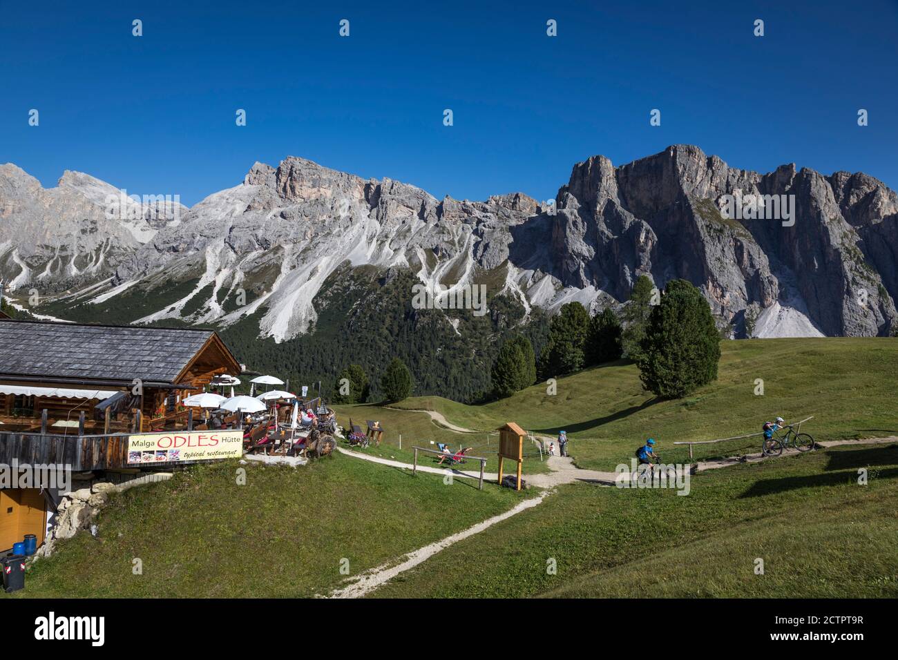 Cyclists leave a mountain restaurant and onto a mountain track with spectacular views of the Dolomite mountains all around , Val Gardena, Italy. Stock Photo