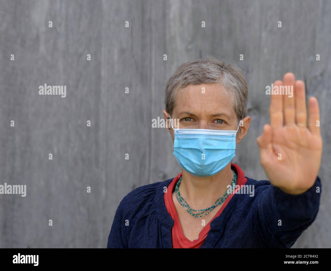 Middle-aged Caucasian woman with short hair wears a blue surgical face mask and makes the hand palm stop sign during the 2019/20 coronavirus pandemic. Stock Photo