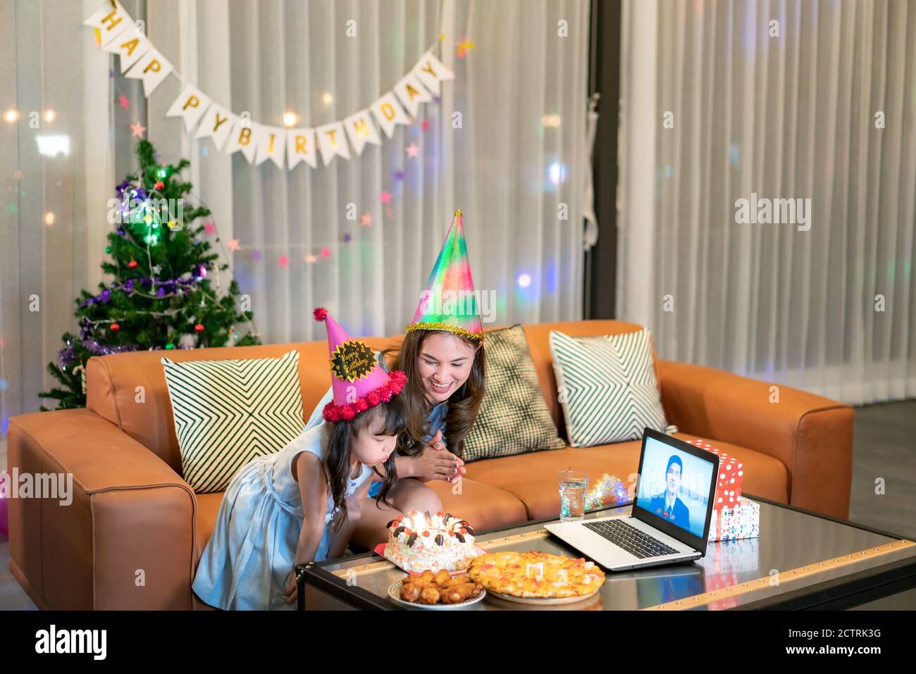 Happy little girl celebrating birthday with her mother at home with father on video call in laptop while blowing candles in cake on table. Stock Photo
