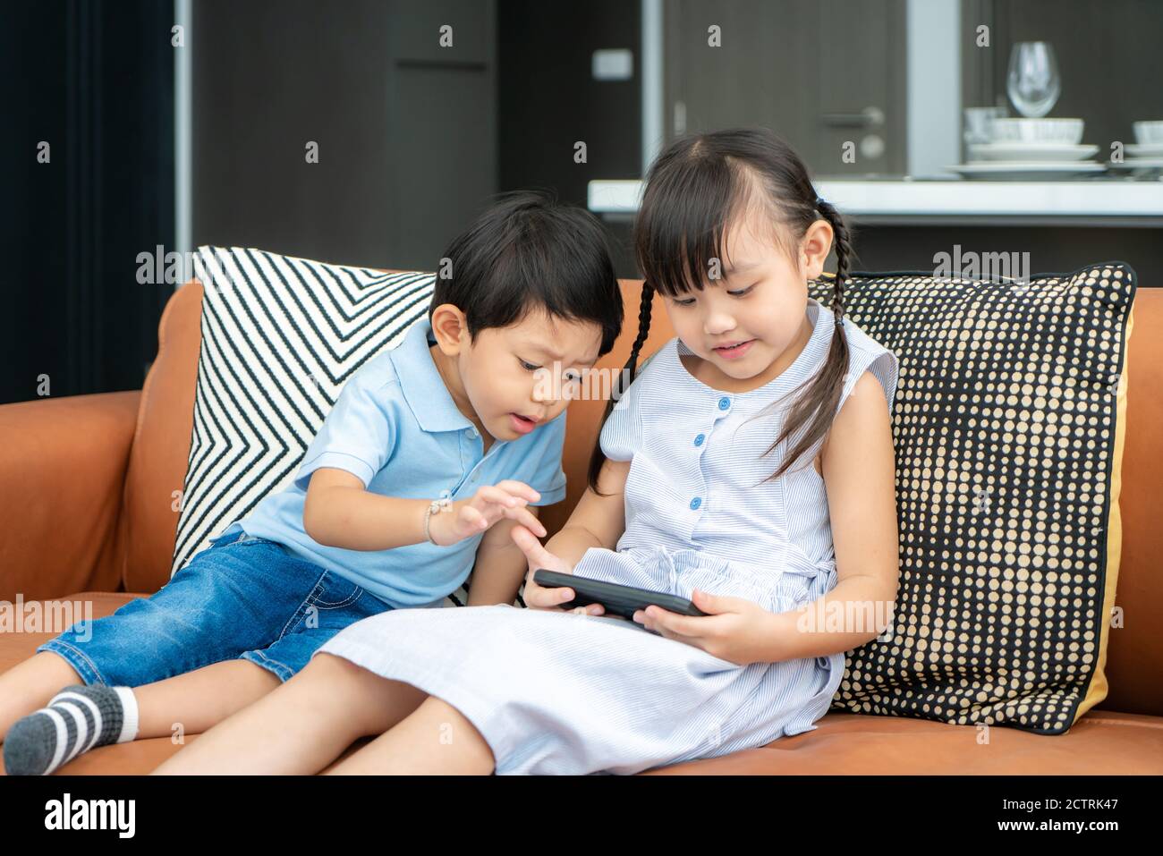 Asian cute sibling child using a smartphone and smiling for watching video or play game togerther while sitting on sofa in living room at home Stock Photo