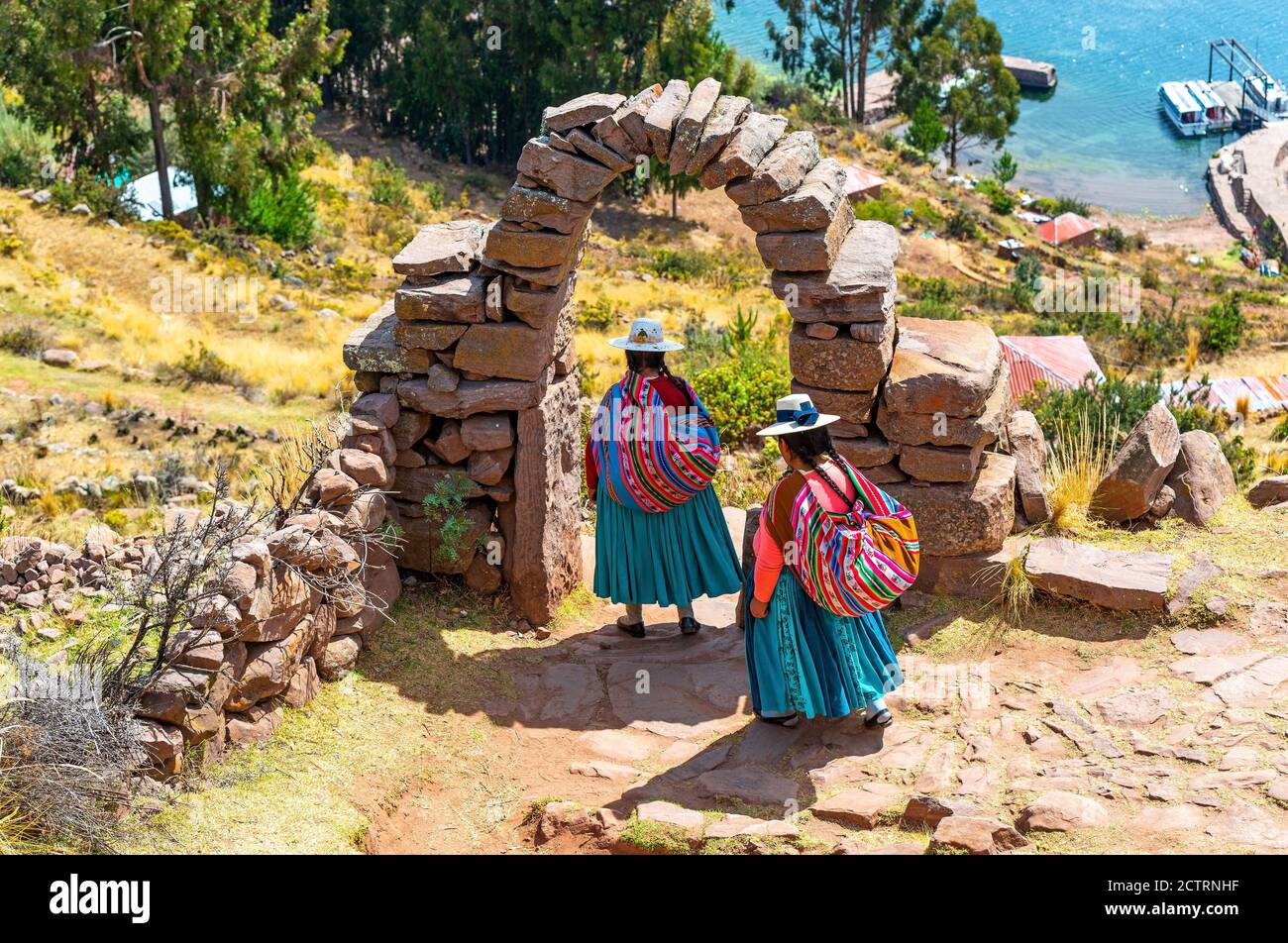 Two peruvian indigenous Quechua women in traditional clothing under an arch walking down steps to a harbor, Taquile island, Titicaca Lake, Peru. Stock Photo