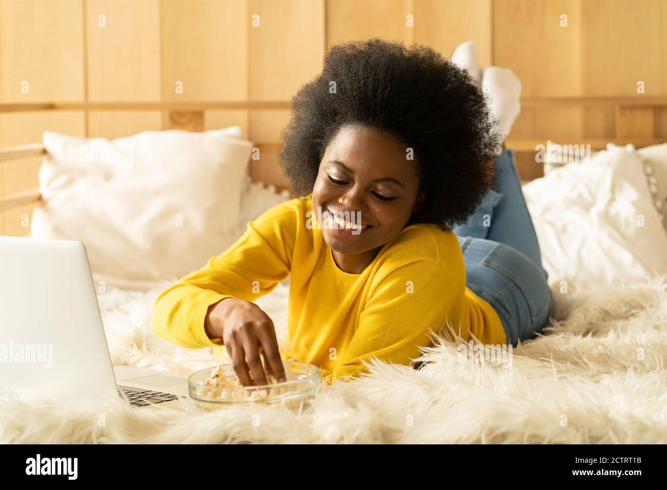 Happy Afro-American woman in yellow sweater relaxing, eating popcorn while watching movie on laptop, talking on video chat, lying in bed. Lazy rest at Stock Photo