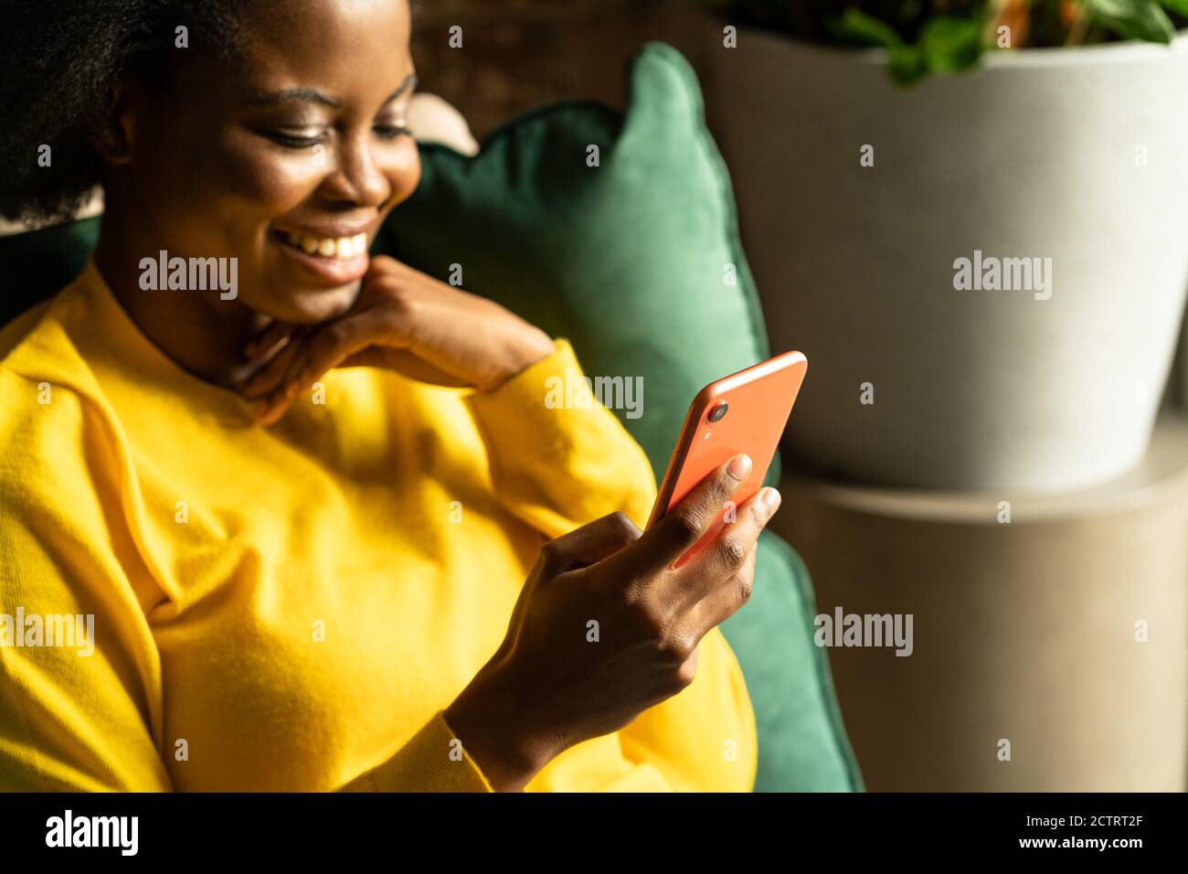 Close up of African American millennial young woman wear yellow sweater sitting in green chair, resting, using smartphone, typing message, taking a br Stock Photo