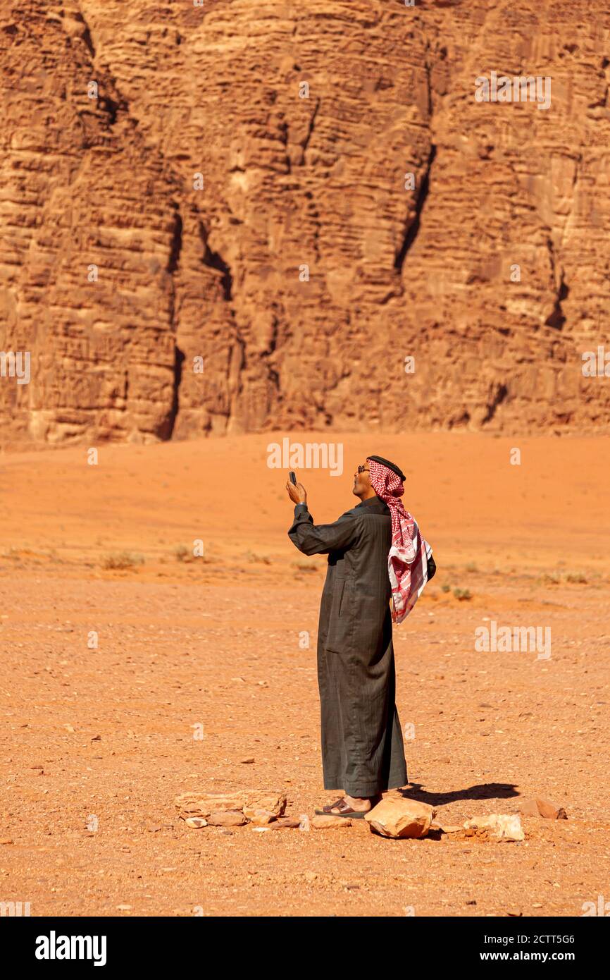 Wadi Rum, Jordan 04/01/2010: An Arabic man wearing traditional keffiyeh, agal, sunglasses and Jubba thobe (long dress) is lifting up his mobile phone Stock Photo