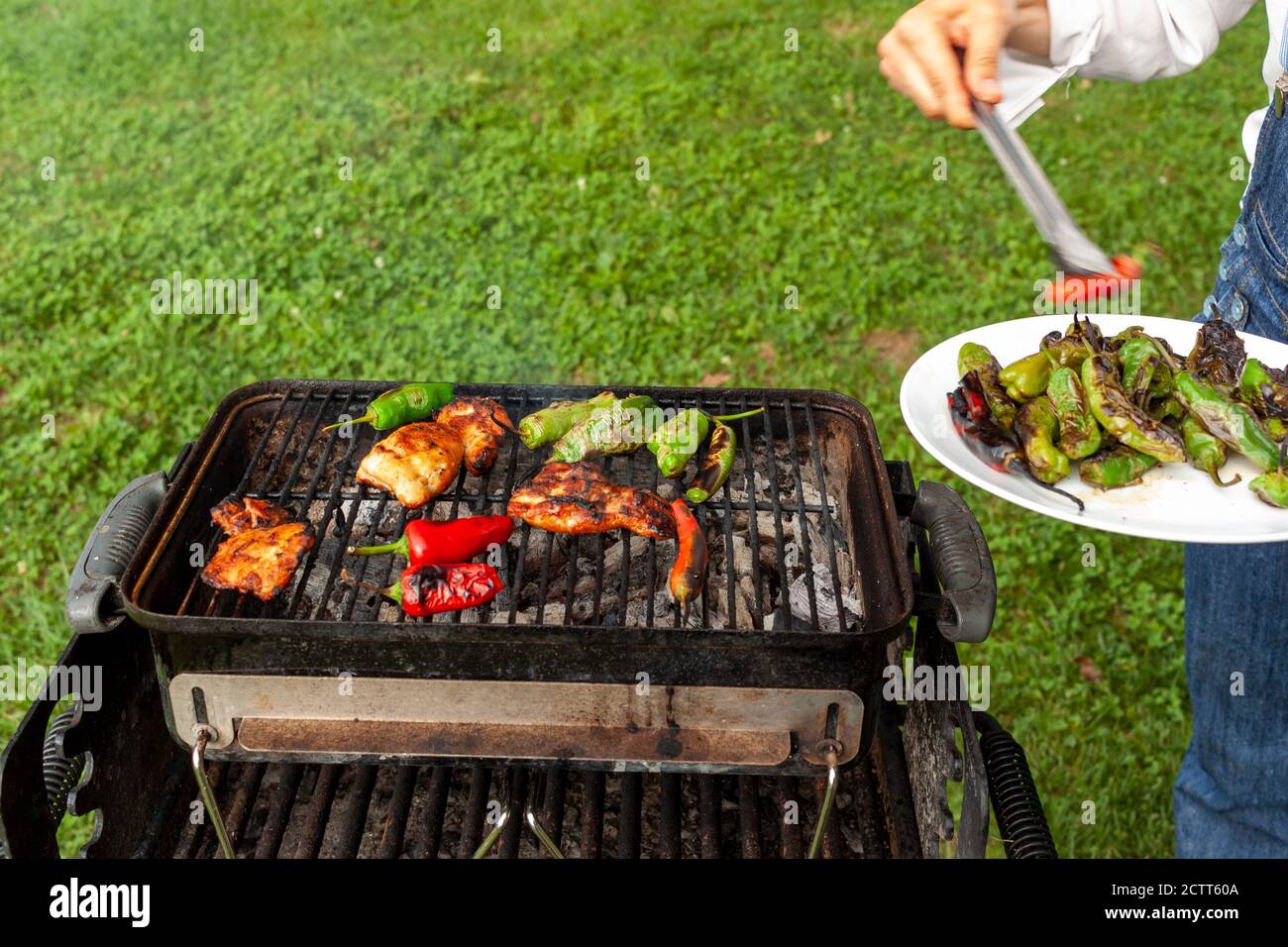 a cheff wearing jean overalls and white shirt is by a charcoal grill where marinated chicken slices, red and green peppers are fire grilled. She uses Stock Photo