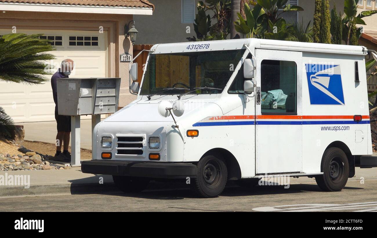 A US Postal Service delivery worker brings mail to a mailbox in suburban neighborhood. He is wearing a mask due to Covid-19 regulations. Stock Photo