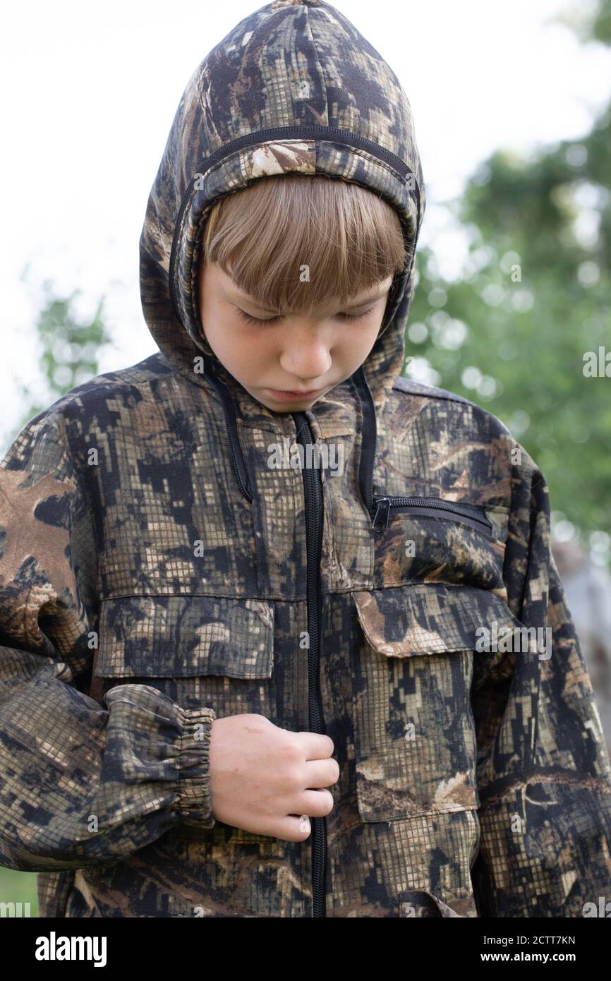 Caucasian boy in masking camouflage uniform saddened outdoor. Plays military or hunter. Stock Photo