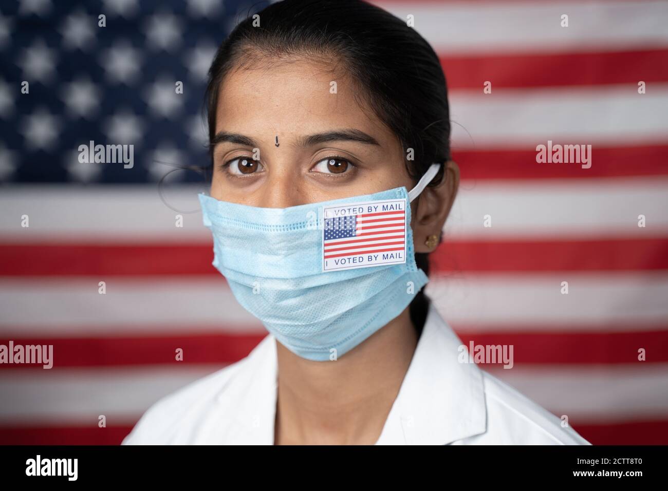 Close up face of Girl with I voted by main sticker on her medical mask with US flag as background - Concept of mail in voting in US election. Stock Photo
