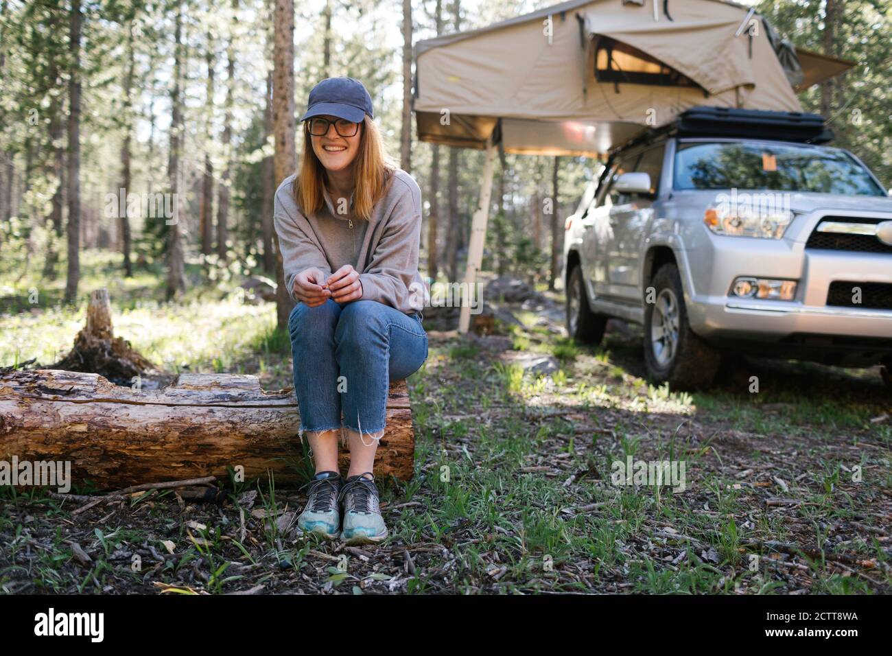 Portrait of smiling woman sitting on log on camping, car with tent in background, Wasatch Cache National Forest Stock Photo