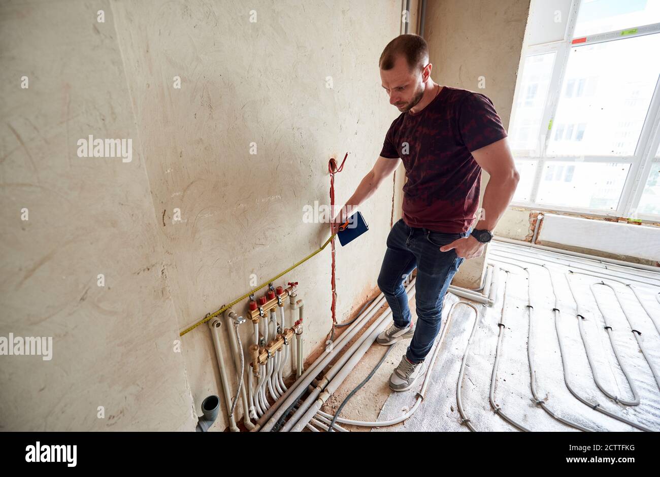 Serious man measuring wall with tape measure in room with underfloor heating pipes. Handyman holding notebook and taking measurements while working on renovation of apartment. Repair concept Stock Photo