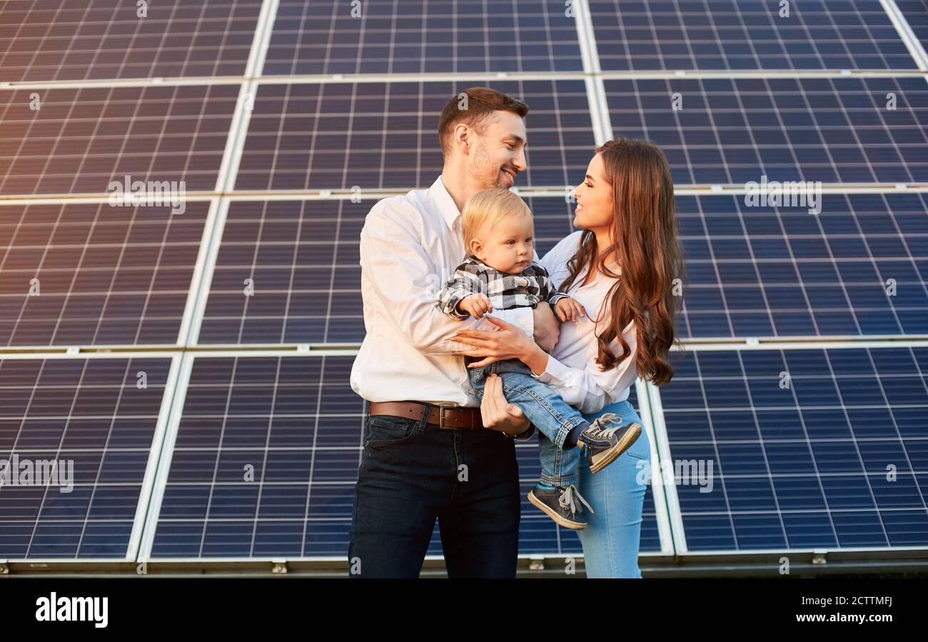 Young family with a small child in her arms on a background of solar panels. A man and a woman look at each other with love. Solar energy concept image Stock Photo