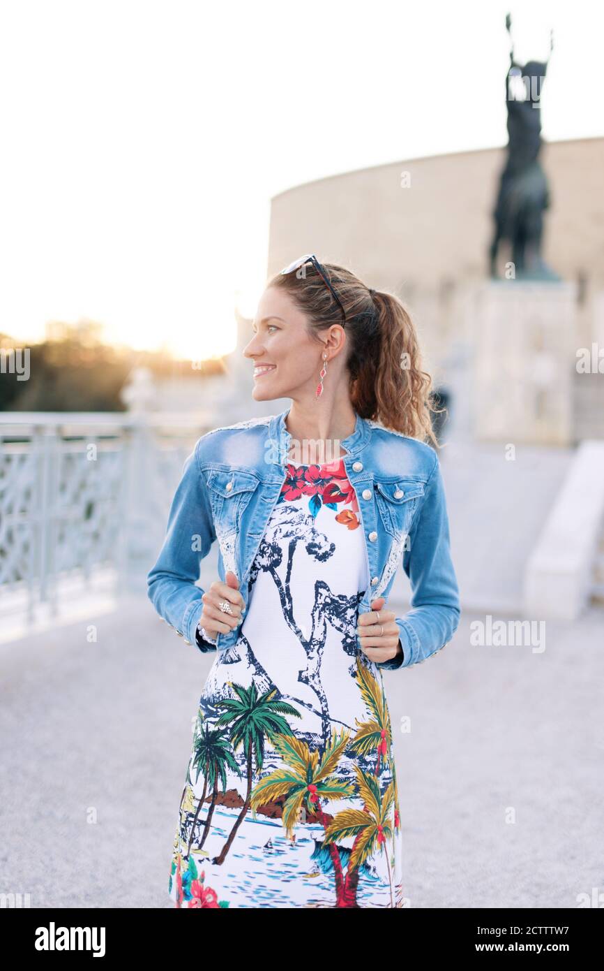 Young cheerful urban woman holding denim jacket at famous place monument in sunset Stock Photo