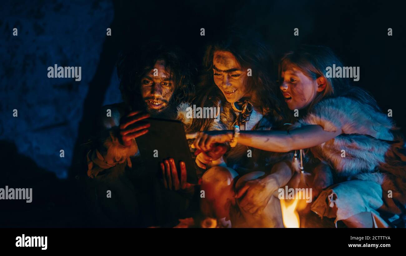 Tribe of Prehistoric, Primitive Hunter-Gatherers Wearing Animal Skins Use Digital Tablet Computer in a Cave at Night. Neanderthal or Homo Sapiens Stock Photo