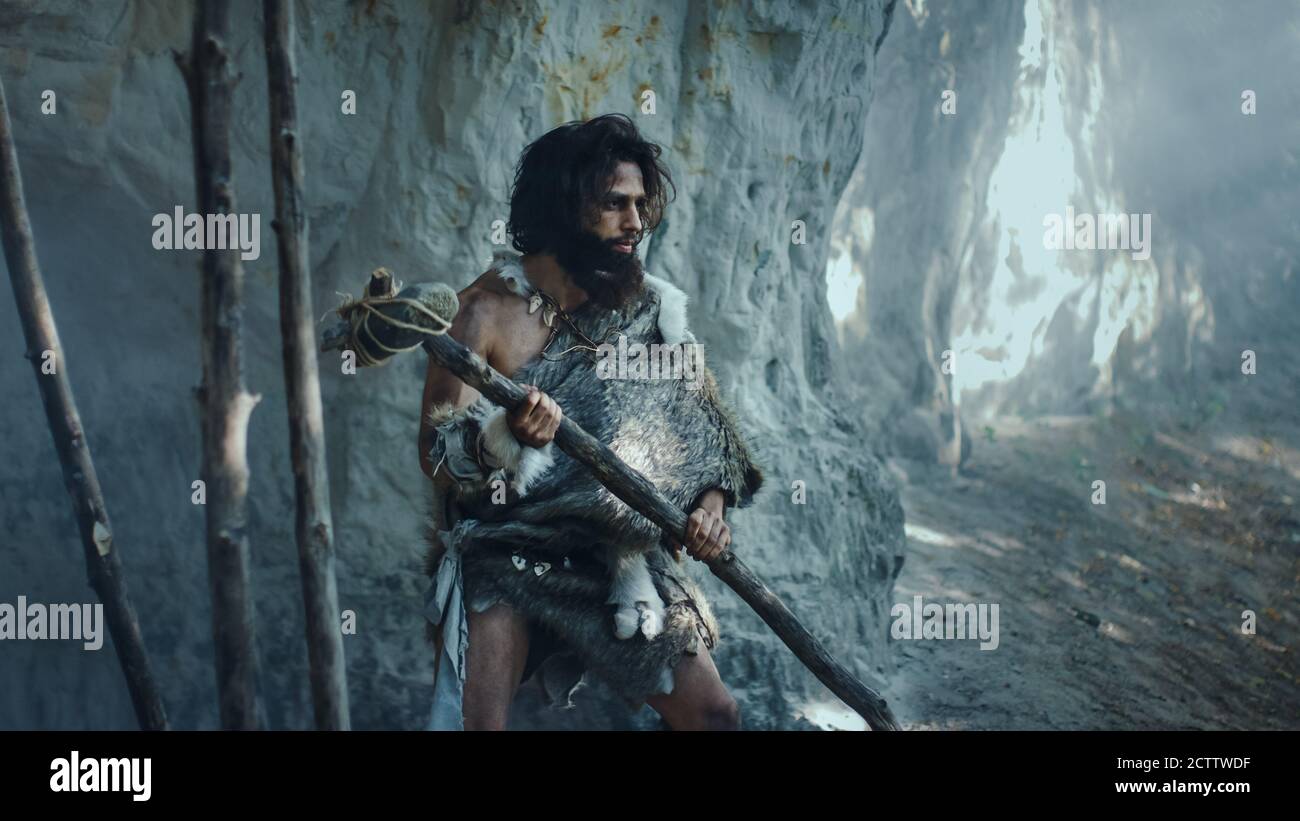 Primeval Caveman Wearing Animal Skin Holds Stone Tipped Hammer Comes out of the Cave and Looks Around Prehistoric Forest, Ready to Hunt Animal Prey Stock Photo