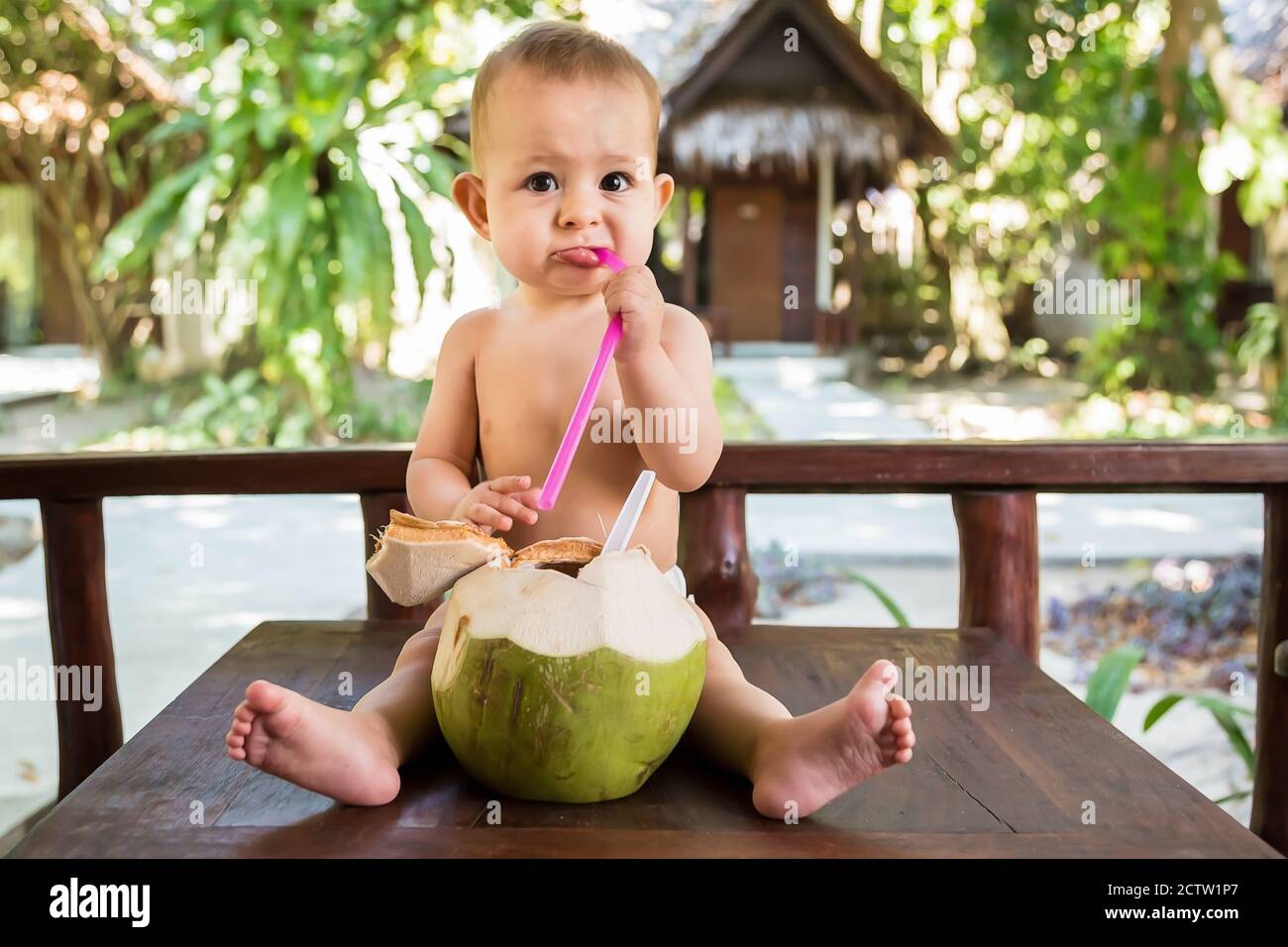 A sad little one year old child sits on a wooden table and drinks coconut milk from fresh green coconut through a straw. Stock Photo