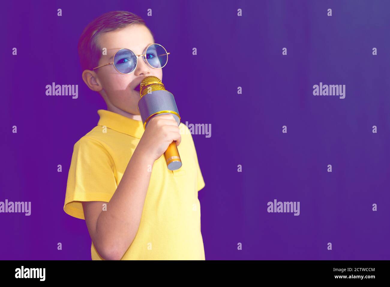 Close up image.The young boy kid child 9s old in yellow t shirt and blue eyeglasses is singing music on purple foggy toned illuminated stage. Stock Photo