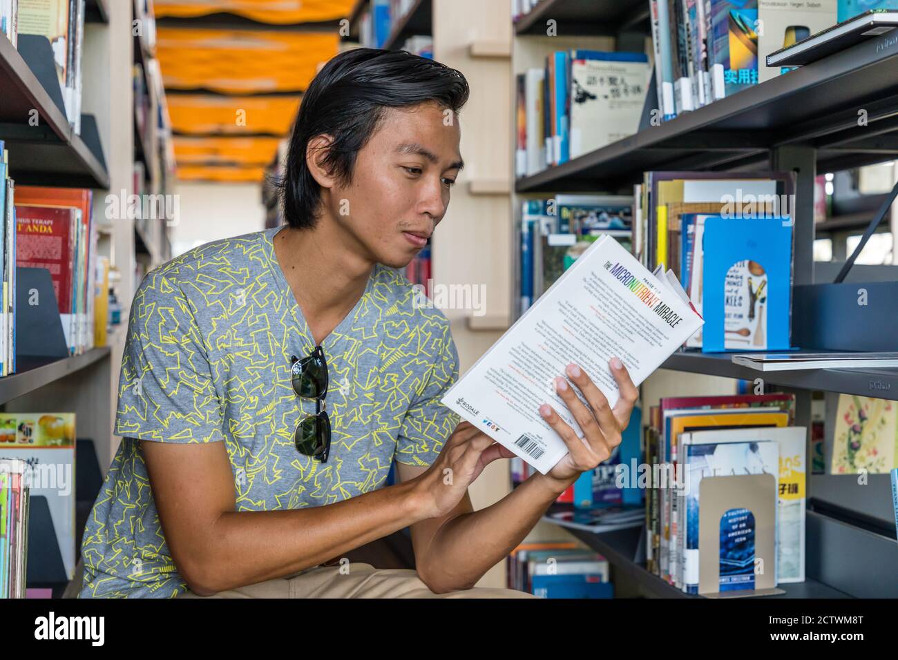 A young man, sitting between the bookshelves and reading a book in the Sabah Regional Library at Tanjung Aru Plaza, Sabah, Kota Kinabalu, Malaysia. Stock Photo