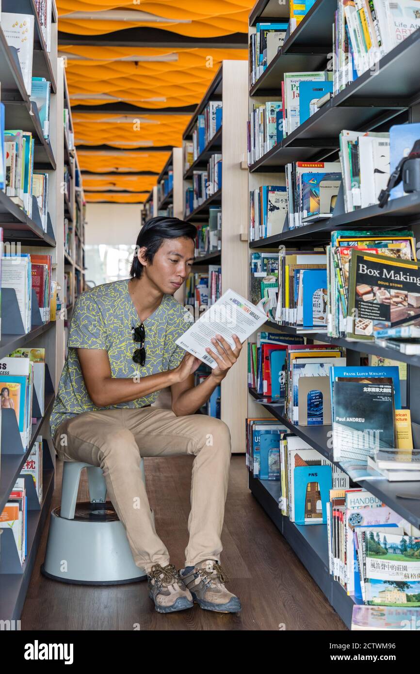 A young man, sitting between the bookshelves and reading a book in the Sabah Regional Library at Tanjung Aru Plaza, Sabah, Kota Kinabalu, Malaysia. Stock Photo