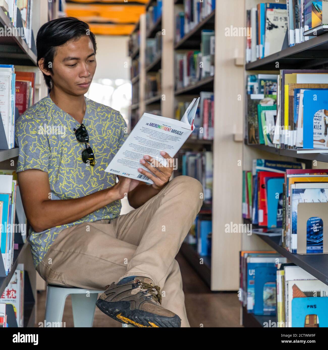 A young man, sitting between the bookshelves and reading a book in the Sabah Regional Library at Tanjung Aru Plaza, Sabah, Kota Kinabalu, Malaysia. Stock Photo