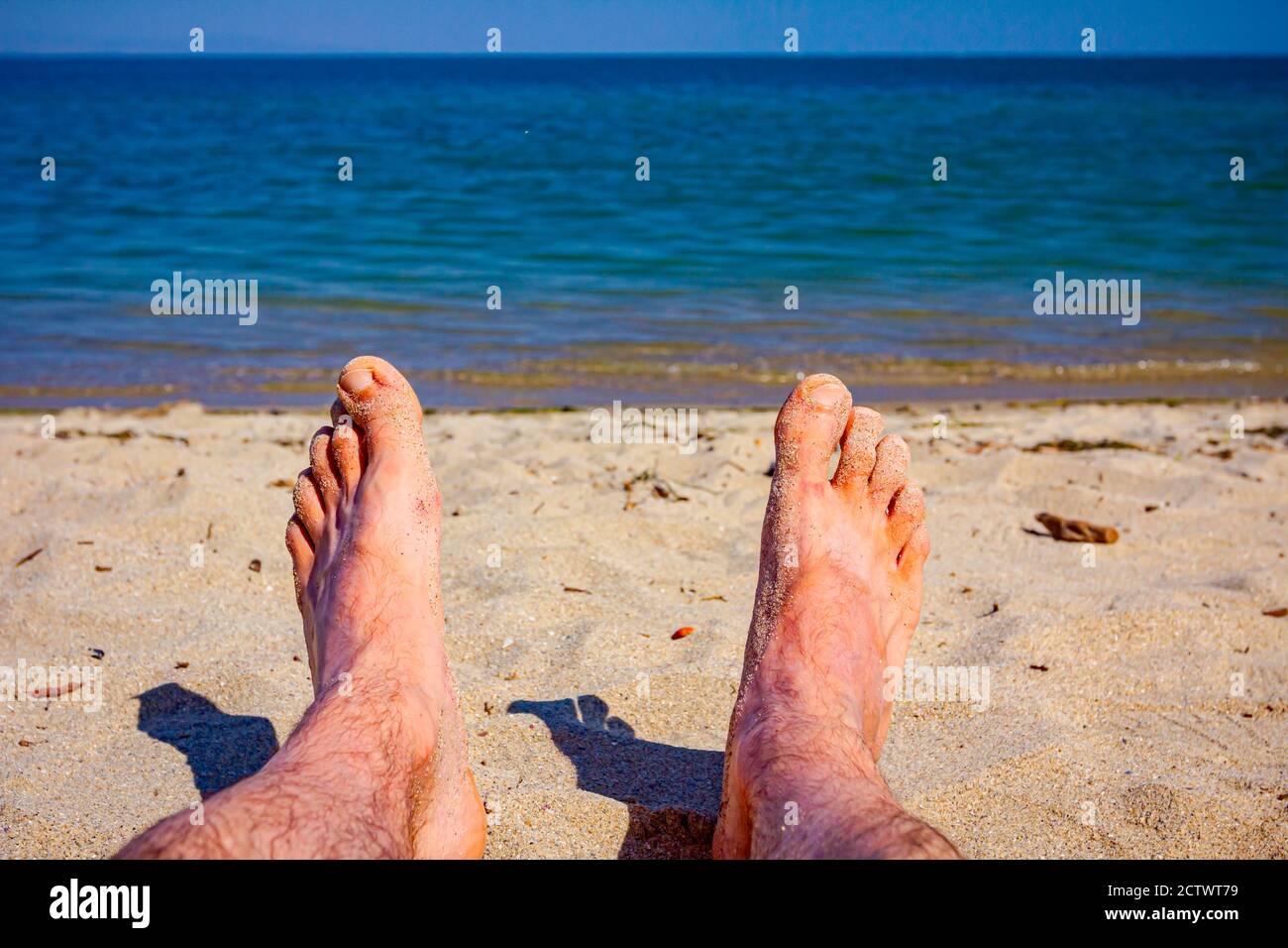 Man's legs are sunbathing by lying carefree on sand next to the coastline, on public beach. Stock Photo