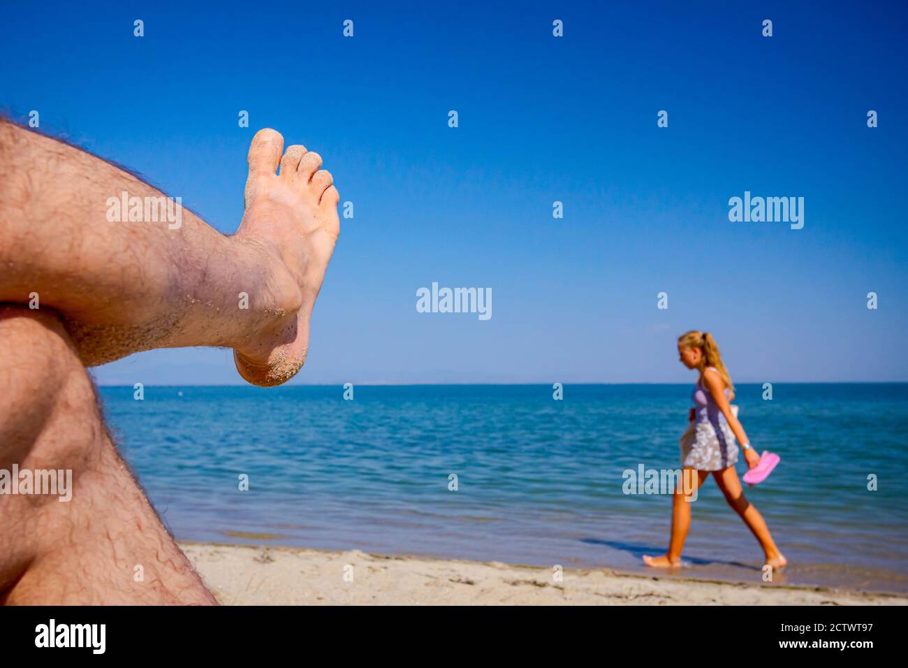 Man's crossed legs are sunbathing by lying carefree on sand next to the coastline, on public beach. Stock Photo