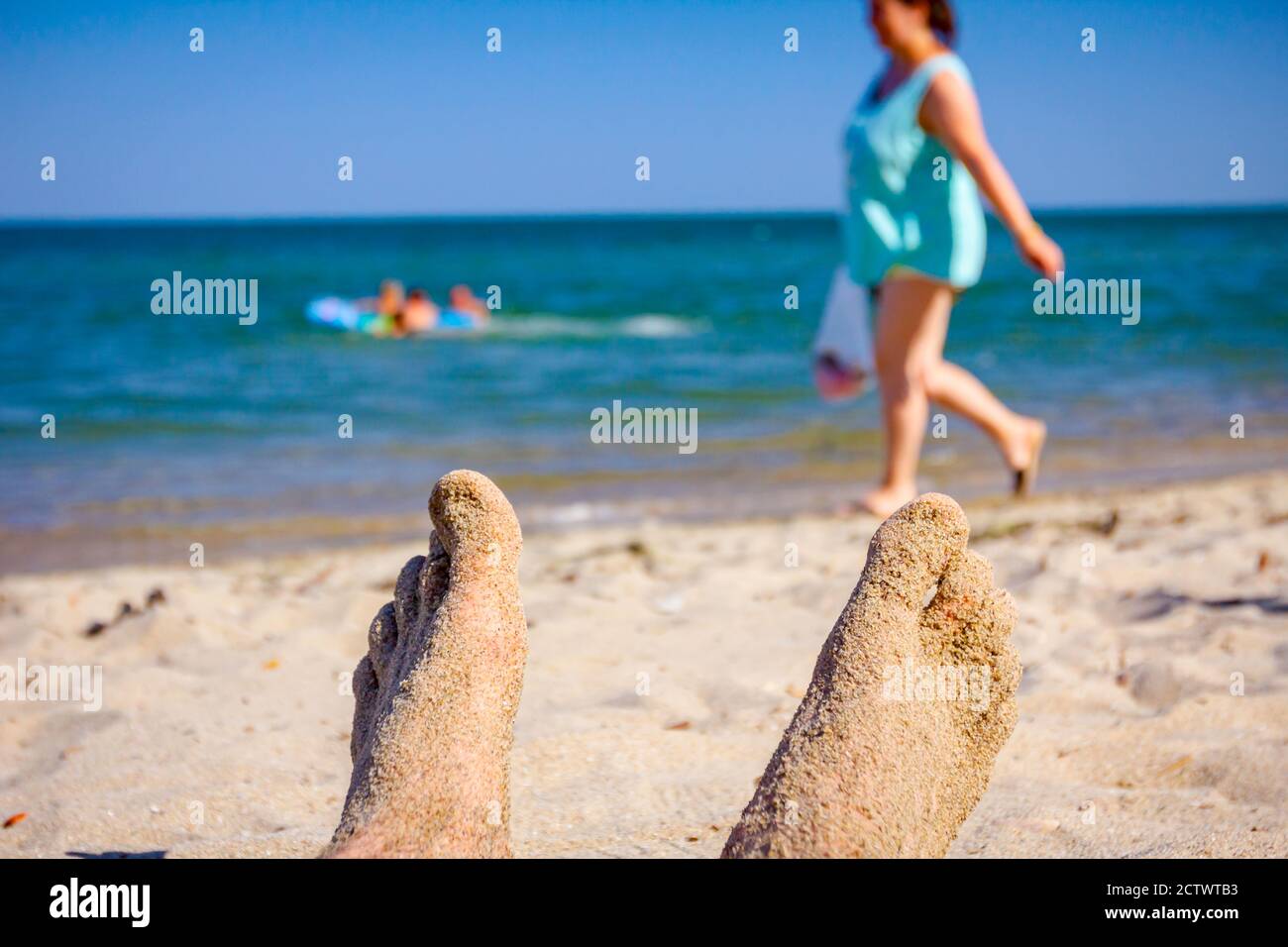 Man's crossed legs are sunbathing by lying carefree on sand next to the coastline, on public beach. Stock Photo
