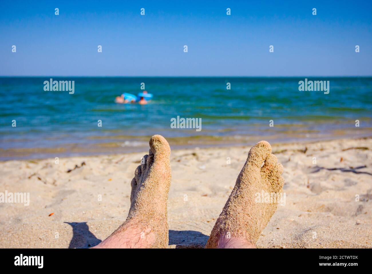 Man's legs are sunbathing by lying carefree on sand next to the coastline, on public beach. Stock Photo
