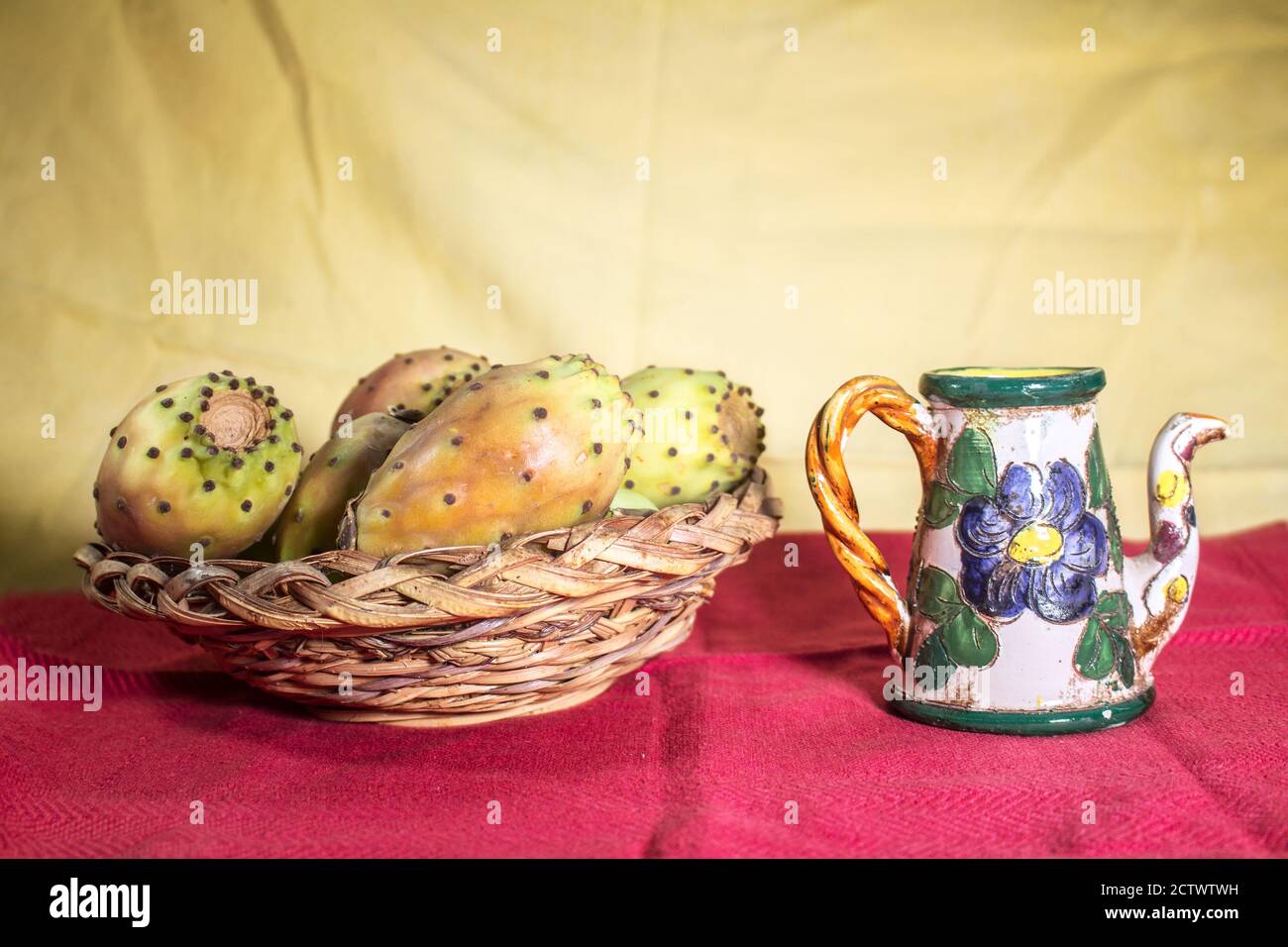 Still life on red surface with wicker basket full of prickly pears and a small decorated ceramic jug Stock Photo