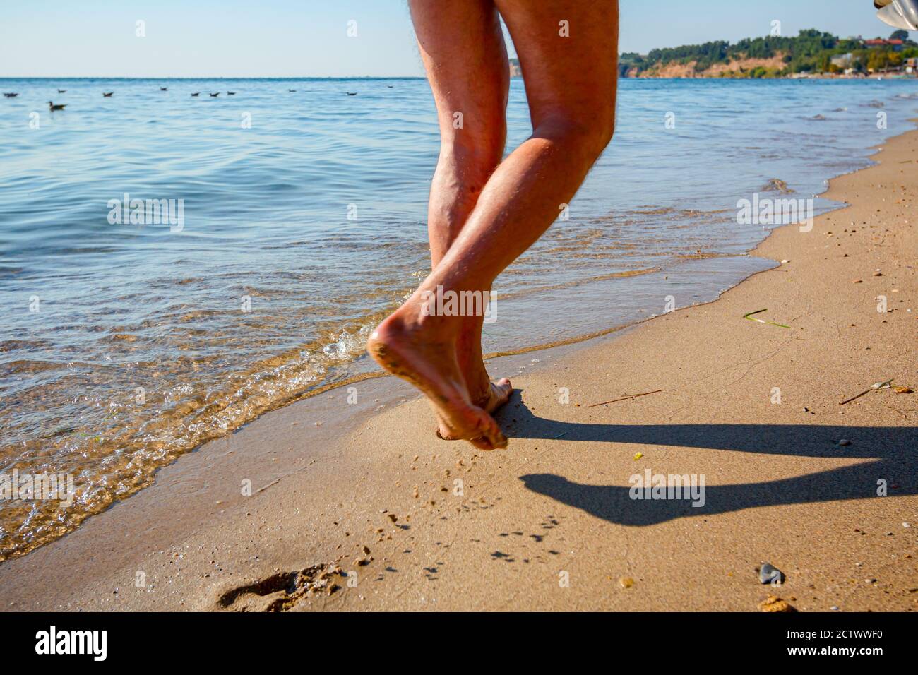 Man's legs are walking barefoot on the sandy beach next to the shallow sea water. Stock Photo