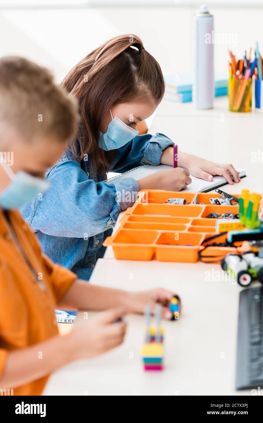 Selective focus of schoolchildren in medical masks modeling robot and writing on notebook in stem school Stock Photo