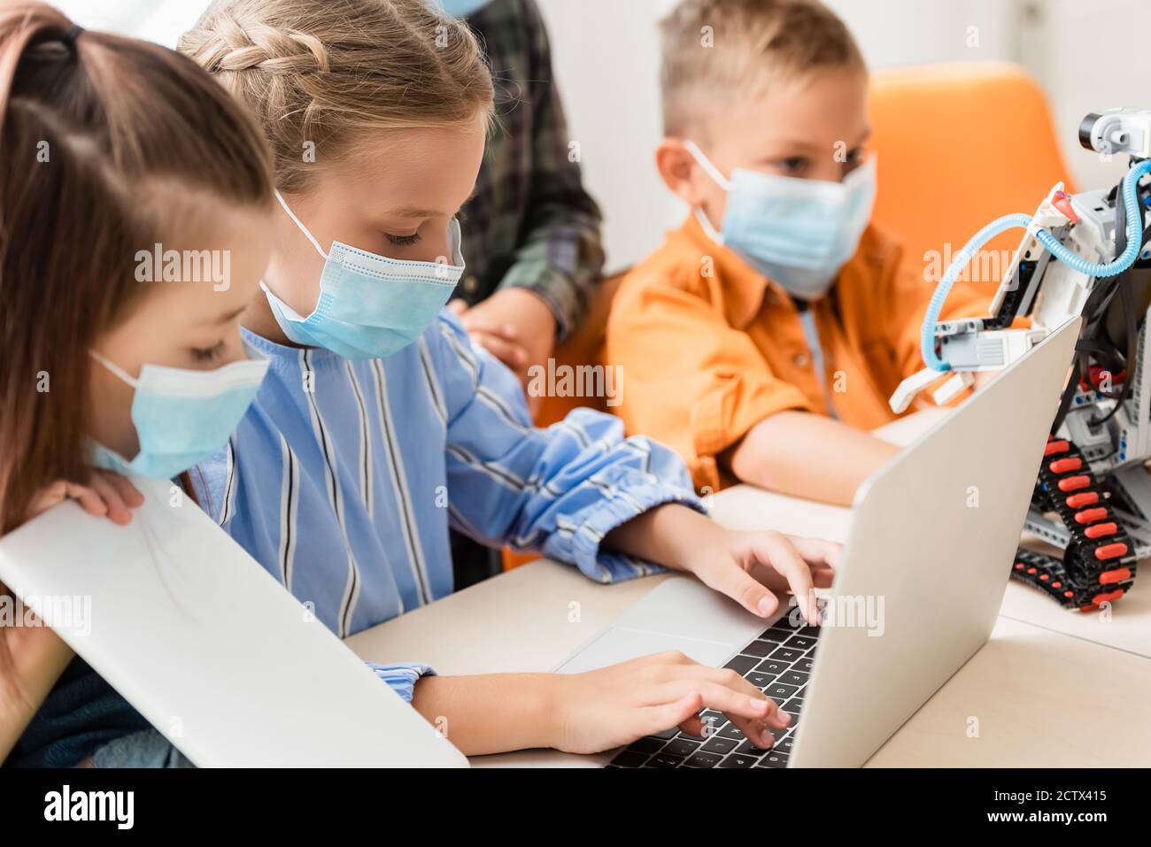 Selective focus of kids in medical masks using laptop and digital tablet near robot in school Stock Photo