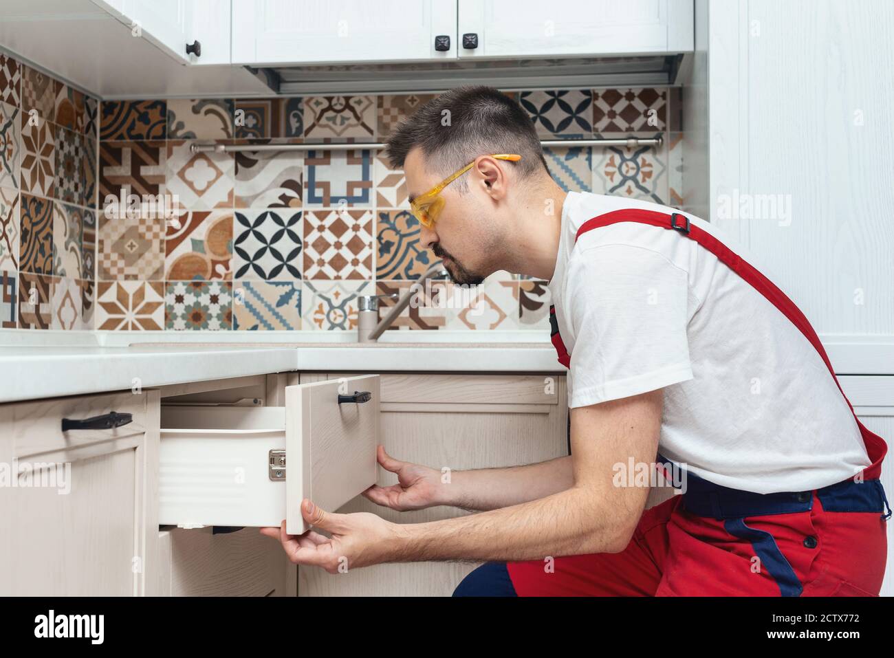 Worker dressed in workers' overall assembing furniture in kitchen. home