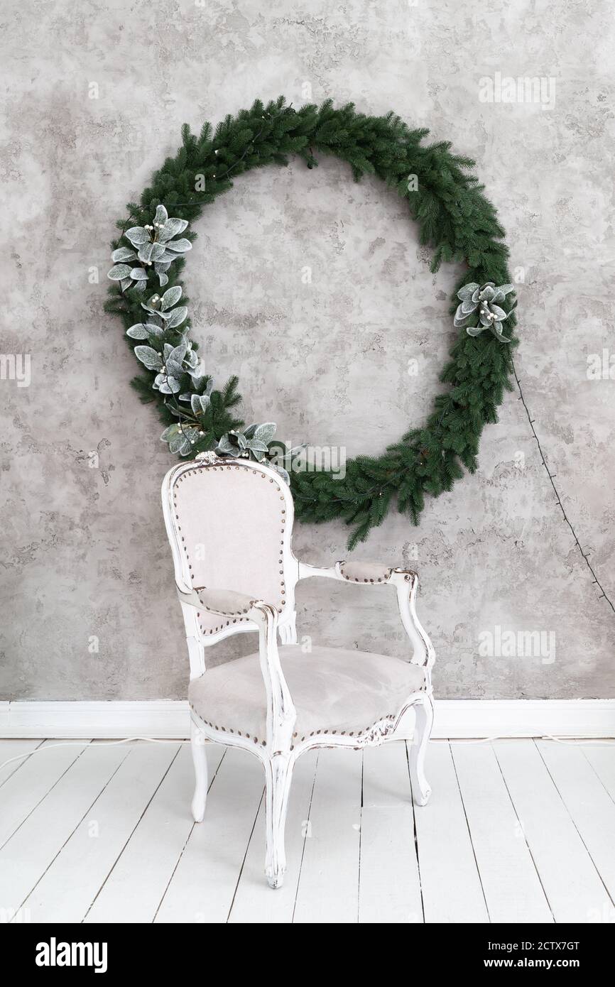 Christmas interior of the room. Gray chair stands under a light wall on which hangs a Christmas wreath. Green conifer wreath decorated with silver Stock Photo