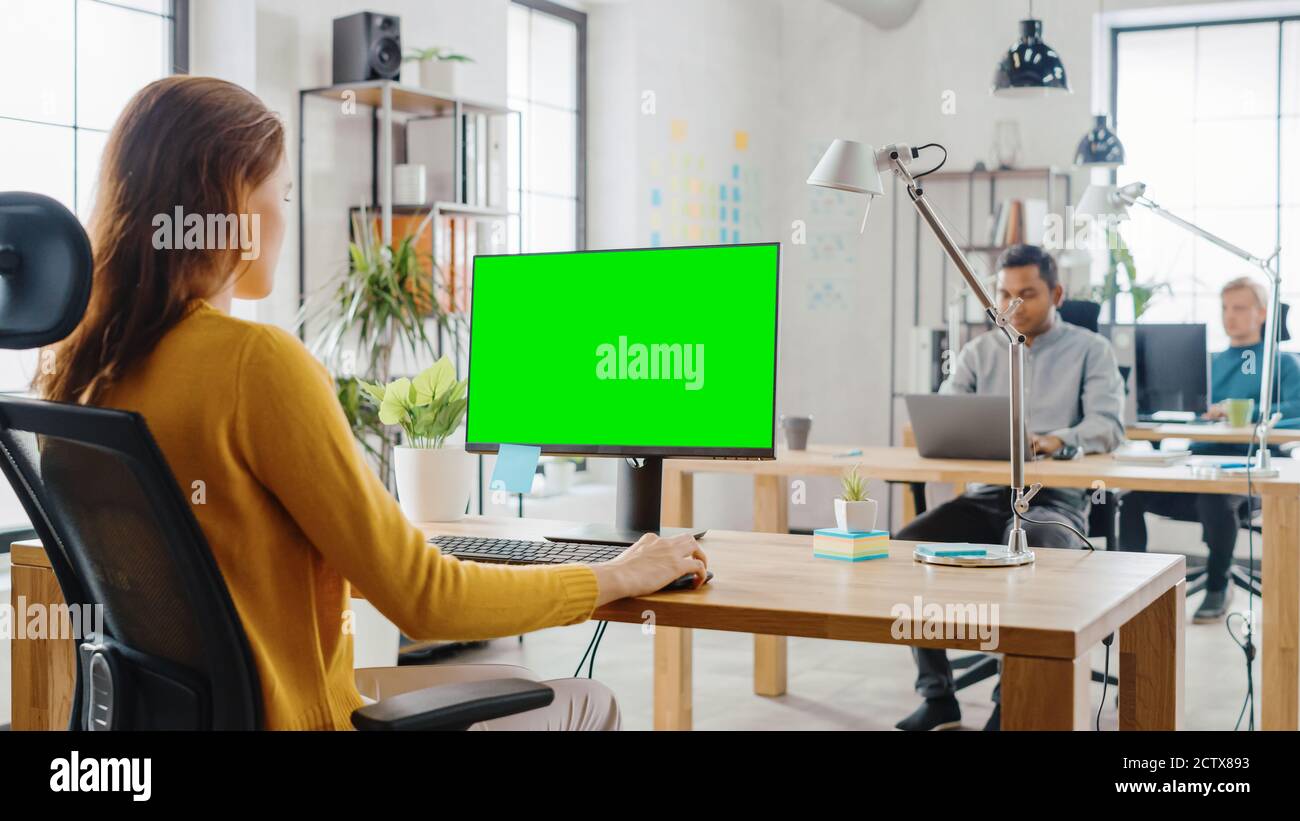 Over the Shoulder: Creative Young Woman Sitting at Her Desk Using Desktop Computer with Mock-up Green Screen. In the Background Bright Office where Stock Photo