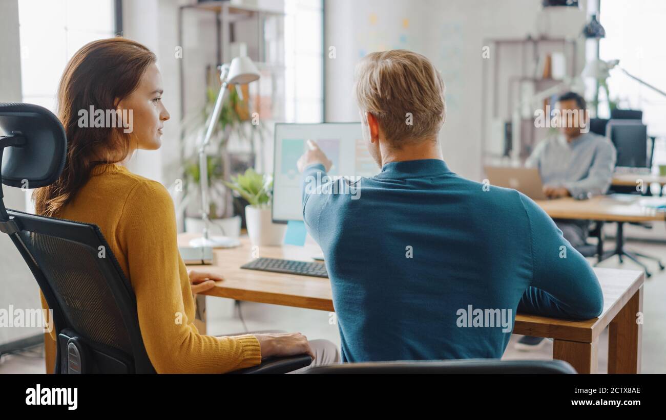 Two Mobile Software Developers Work on Desktop Computer, They Discuss Smartphone Application Design. In the Background Modern Stylish Office with Stock Photo