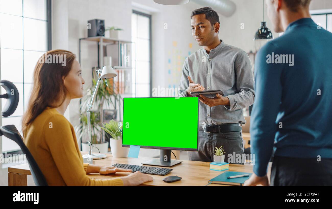 Three Start-up Entrepreneurs Work on Desktop Computer with Green Mock-up Screen, They Discuss Project Design, Use Touch Screen Digital Tablet. Modern Stock Photo