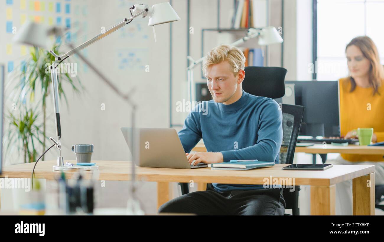 Handsome Caucasian IT Specialist Sitting at His Desk Works on a Laptop under His Business Project. Diverse Team of Young Professionals Working in Stock Photo