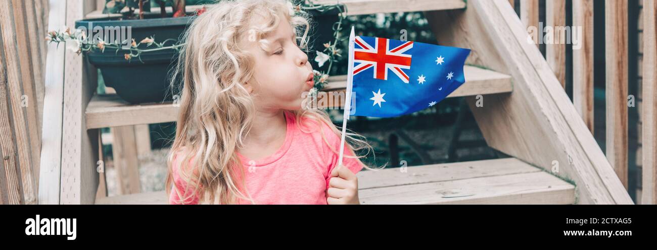 Happy Caucasian girl holding blowing on Australian flag. Funny child sitting on backyard at home and holding Australia flag. Kid citizen celebrating Stock Photo