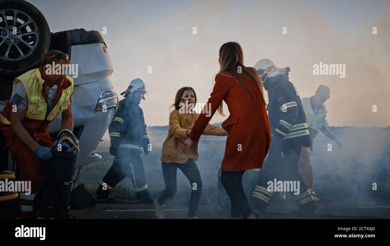 Car Crash Traffic Accident: Injured Young Girl Reunites with Her Loving Mother. In the Background Rollover Car and Courageous Paramedics and Firemen Stock Photo