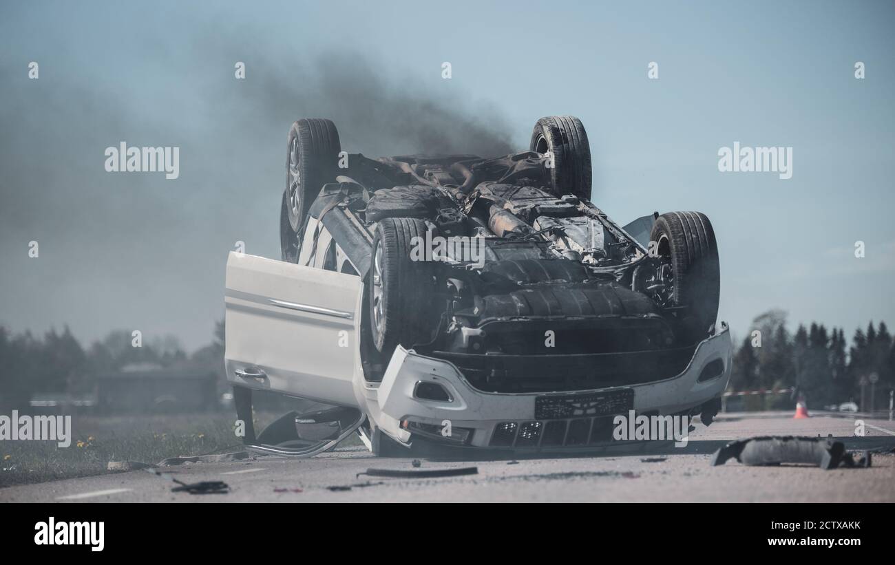 Horrific Traffic Accident Rollover Smoking and Burning Vehicle Lying on its Roof in the Middle of the Road after Collision. Daytime Crash Scene with Stock Photo