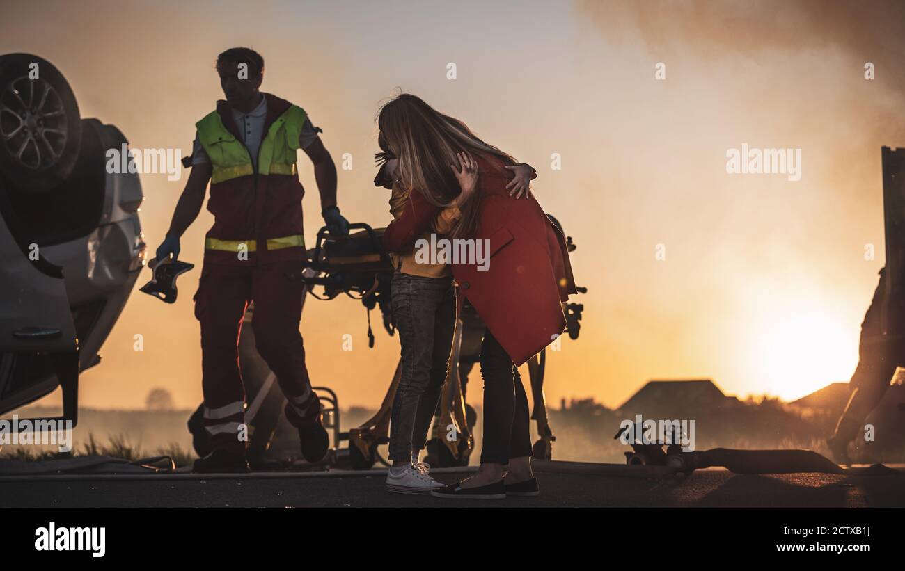 Car Crash Traffic Accident: Injured Young Girl Reunites with Her Loving Mother. In the Background Rollover Car and Courageous Paramedics and Firemen Stock Photo