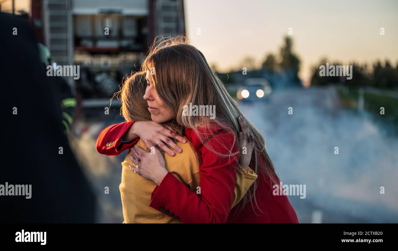 Car Crash Traffic Accident: Injured Young Girl Reunites with Her Loving Mother. In the Background Fire engine and Courageous Paramedics and Firemen Stock Photo