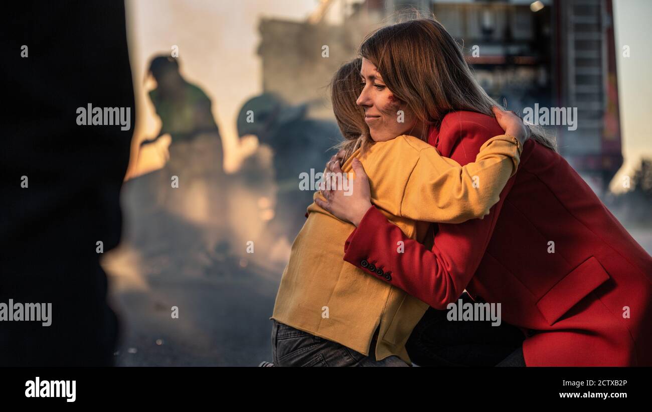 Car Crash Traffic Accident: Injured Young Girl Reunites with Her Loving Mother. In the Background Fire engine and Courageous Paramedics and Firemen Stock Photo