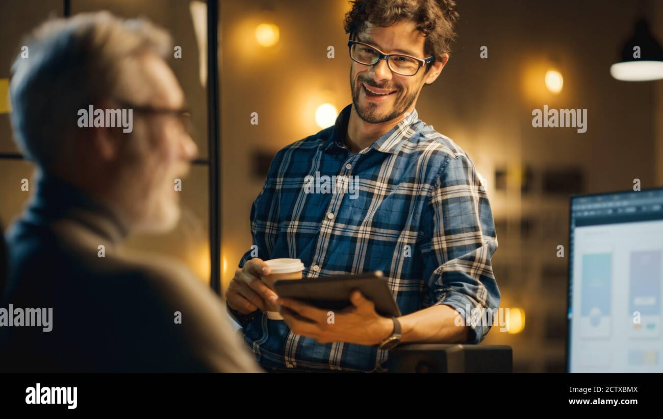 In the Evening Creative Middle Aged Smartphone Software Developer Works on a Desktop Computer with Screen Showing Application Design, Has Conversation Stock Photo